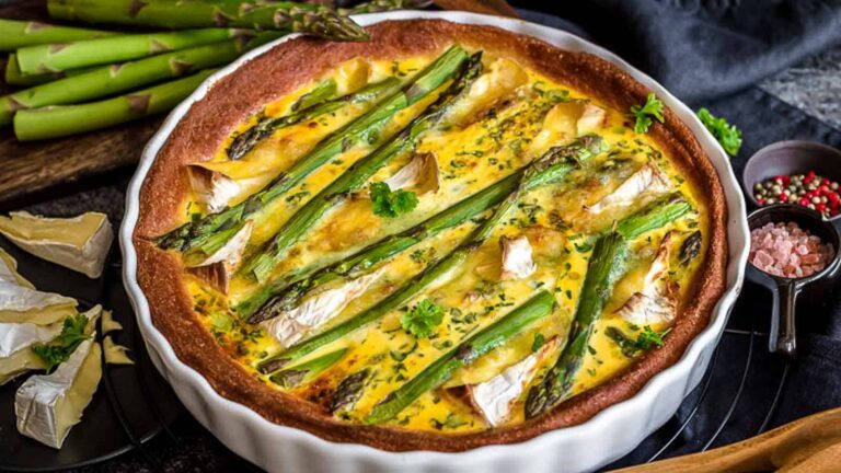 A baked quiche topped with asparagus spears and herbs sits in a white pie dish, with raw asparagus, cheese, and seasonings nearby.