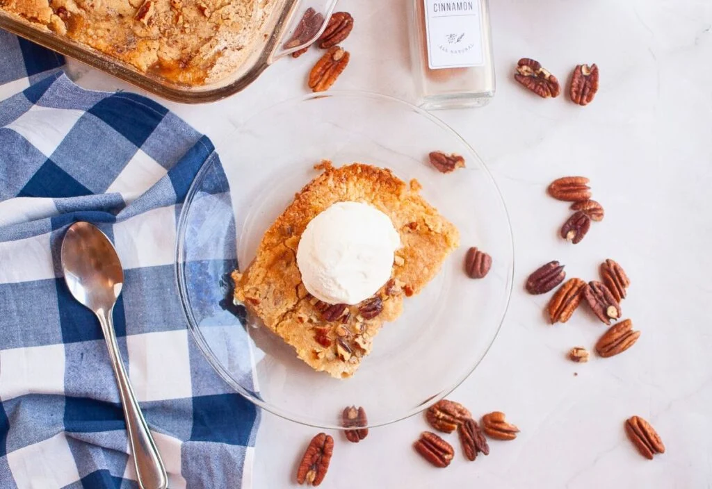 A slice of Autumn Bliss Apple Cake topped with vanilla ice cream sits on a glass plate, surrounded by pecans, a spoon, a blue checkered cloth, and a cinnamon jar.