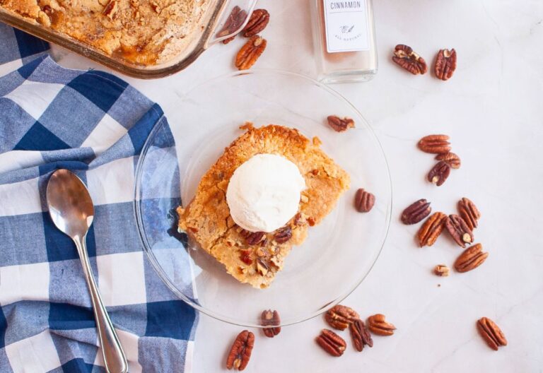 A slice of Autumn Bliss Apple Cake topped with vanilla ice cream sits on a glass plate, surrounded by pecans, a spoon, a blue checkered cloth, and a cinnamon jar.