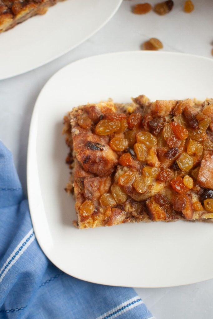A slice of cinnamon roll brunch bake topped with golden raisins sits on a white plate next to a blue napkin.