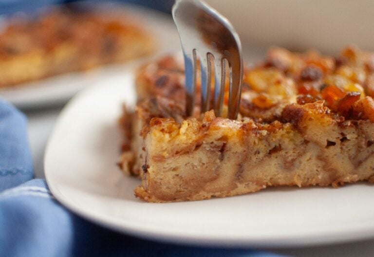 A close-up of a fork cutting into a slice of cinnamon roll brunch bake on a white plate.