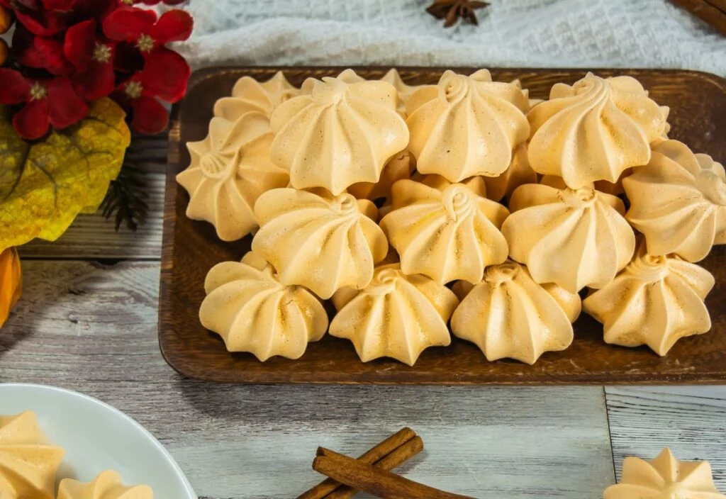 A wooden tray filled with light orange pumpkin meringues arranged in rows on a rustic wooden table, with decorative flowers and cinnamon sticks nearby.