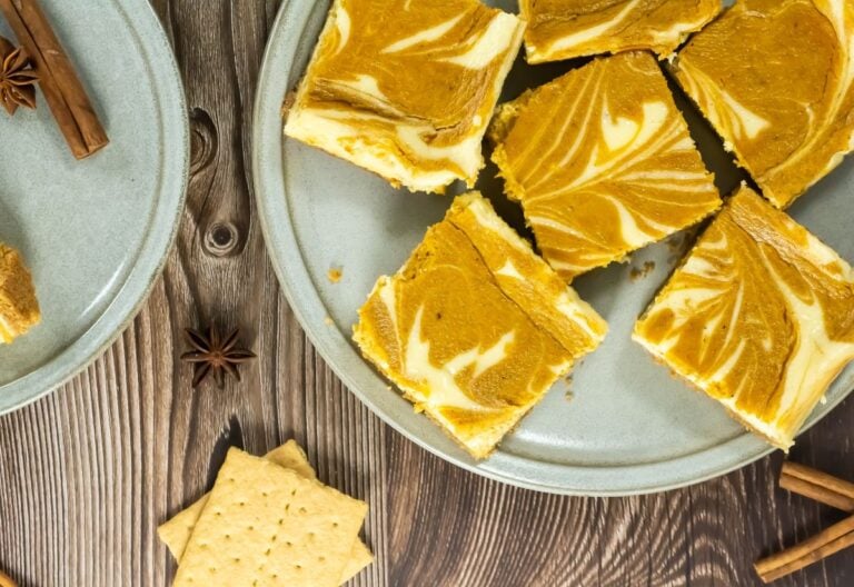 Six pumpkin cheesecake bars with marbled tops are arranged on a gray plate, next to graham crackers, cinnamon sticks, and a star anise on a wooden surface.
