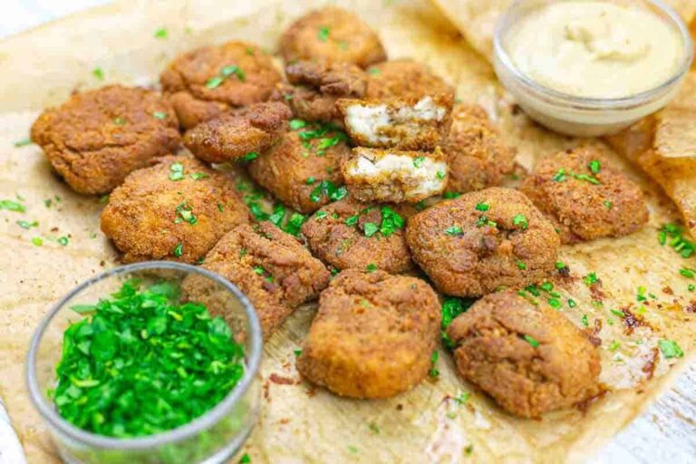 A batch of golden-brown vegan chicken nuggets garnished with chopped herbs, served on parchment paper with bowls of dipping sauce and chopped greens.