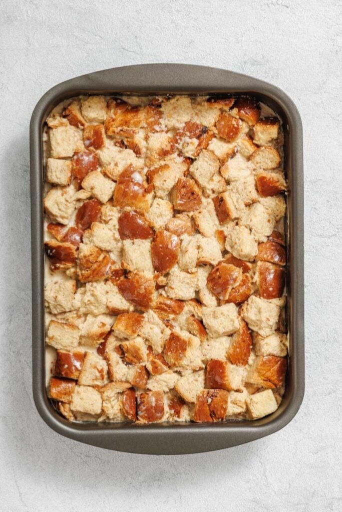 A baking pan filled with cubed bread pieces soaked in a liquid mixture, prepared for baking bread pudding.