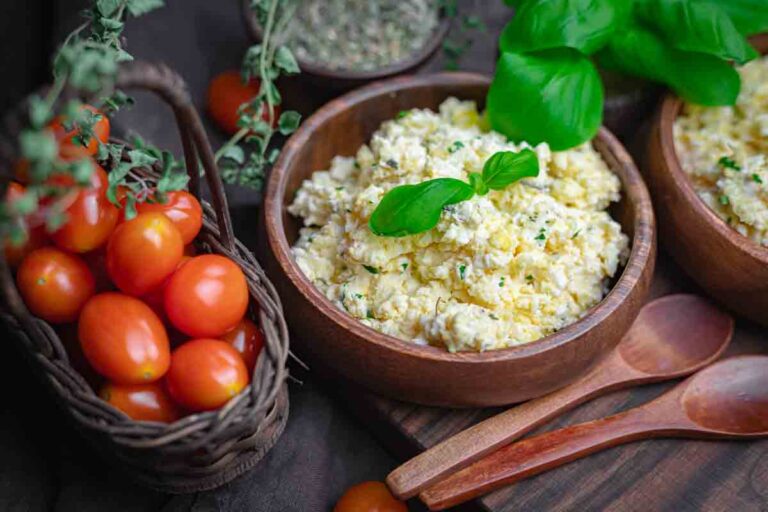 A wooden bowl of scrambled eggs garnished with fresh basil sits next to a basket of cherry tomatoes and two wooden spoons.