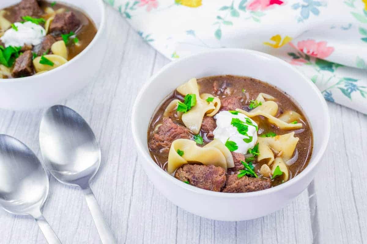 Bowl of beef stroganoff soup with sour cream and parsley garnish on a light wooden table next to two spoons and a floral napkin.