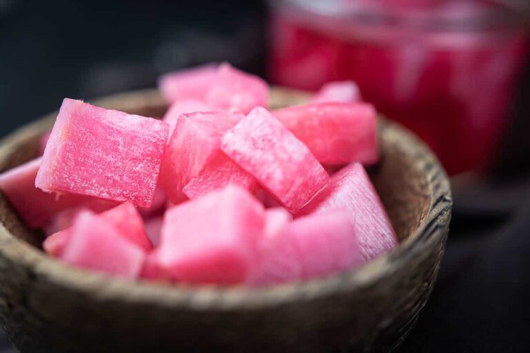 A wooden bowl filled with pink cubes of pickled turnip, with a blurred glass container in the background.