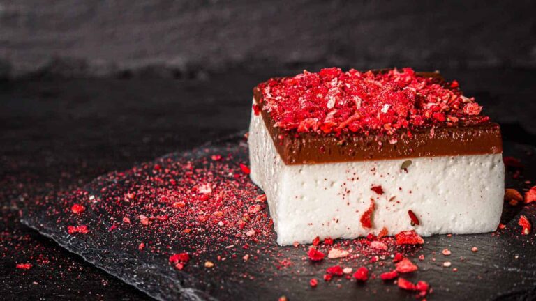 A square piece of dessert with a white base, a chocolate layer on top, and crushed red topping, served on a dark slate surface.