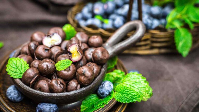 A bowl of chocolate-covered blueberries garnished with fresh mint leaves, with loose blueberries and more mint leaves around the bowl.