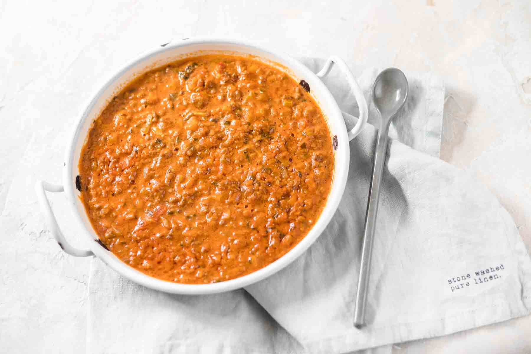 A round white dish filled with Bolognese ragu is placed on a gray cloth.