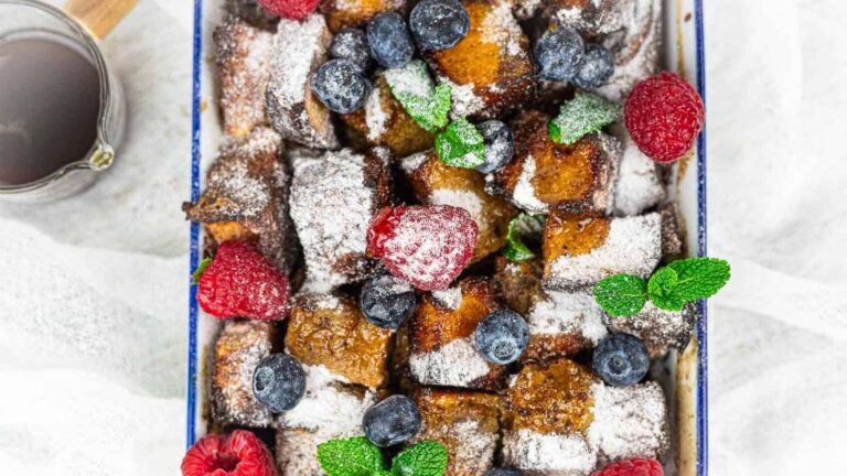 A tray of powdered sugar-dusted French toast bites topped with fresh raspberries, blueberries, and mint leaves.