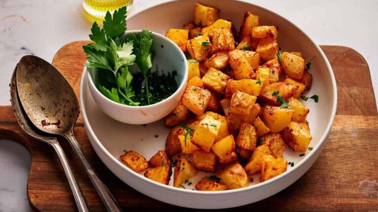 A plate of roasted diced potatoes garnished with chopped herbs, served with a small bowl of fresh parsley and chives on a wooden board with two spoons.