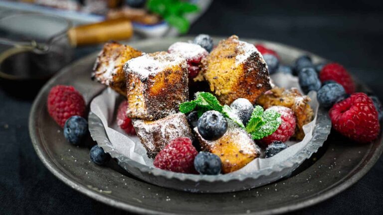 A plate of powdered French toast cubes garnished with fresh raspberries, blueberries, and mint leaves, served on a dark plate.