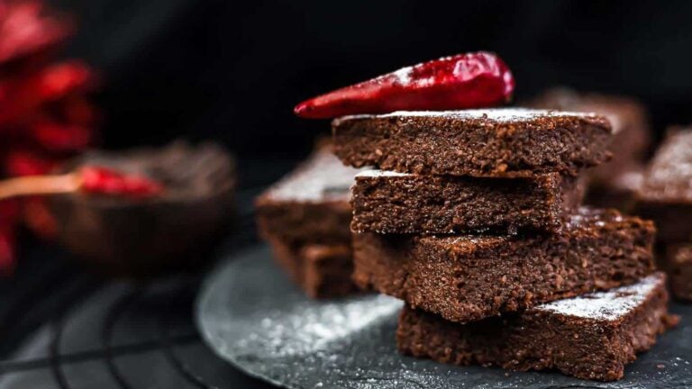 A stack of chocolate brownies dusted with powdered sugar, topped with a whole red chili pepper, on a dark plate.