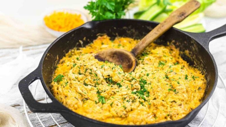 A cast iron skillet filled with creamy, shredded chicken pasta topped with chopped herbs; a wooden spoon rests inside, with ingredients visible in the background.
