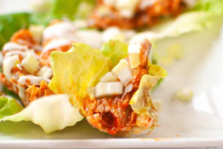 A close-up of a lettuce wrap filled with shredded meat, diced onions, and drizzled with a creamy sauce on a white plate.