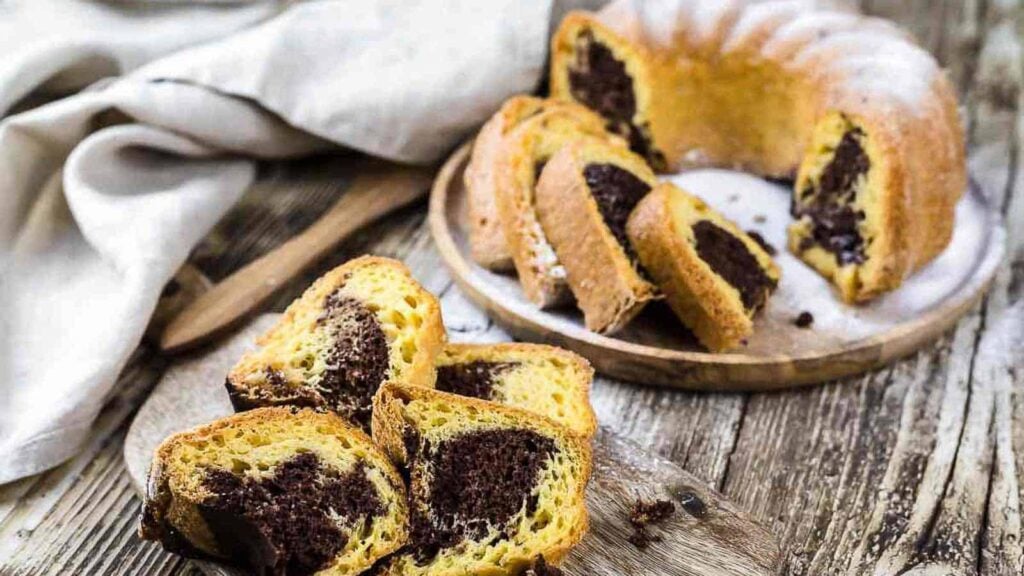 Sliced marble cake with chocolate and vanilla swirls arranged on a wooden board and plate, with a napkin and wooden spoon nearby.