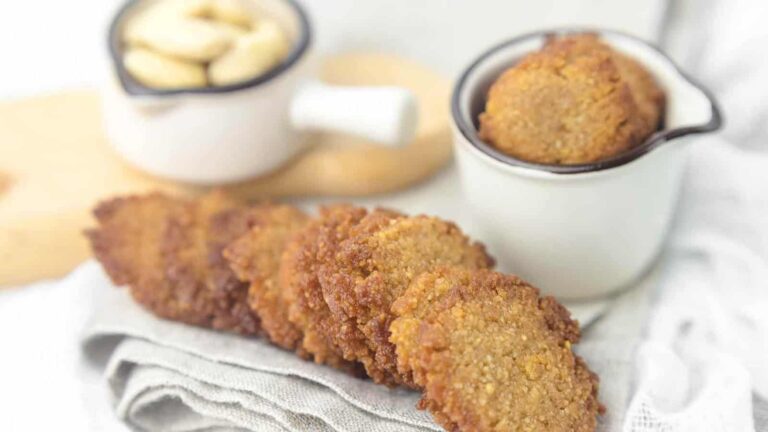 Several golden-brown cookies are arranged on a folded cloth, with more cookies in a small bowl and a bowl of cashews in the background.