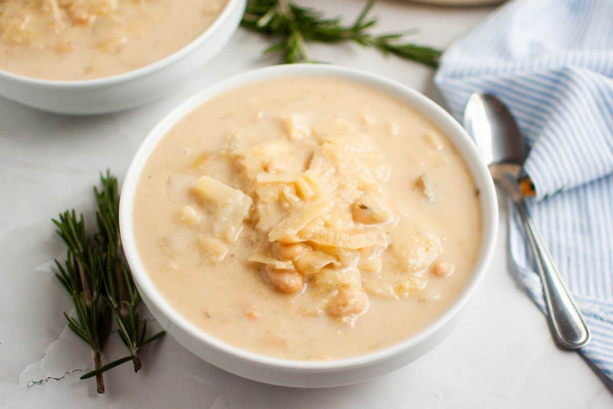 A bowl of Cabbage and Bean Soup with visible chunks of vegetables and beans, garnished with sprigs of rosemary. A spoon and striped napkin are beside the bowl.