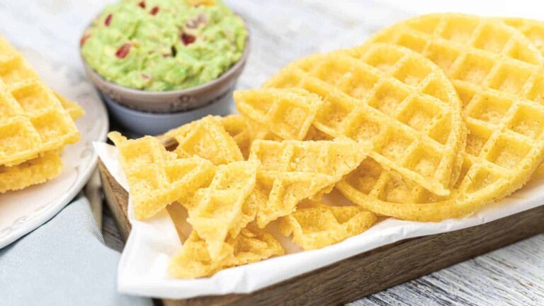 A tray of crispy waffle pieces next to a bowl of guacamole on a white surface.