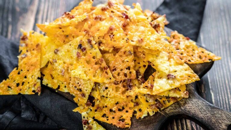 A pile of crispy baked cheese crisps with a golden brown color and specks of seasoning, arranged on a wooden serving board over a dark cloth.