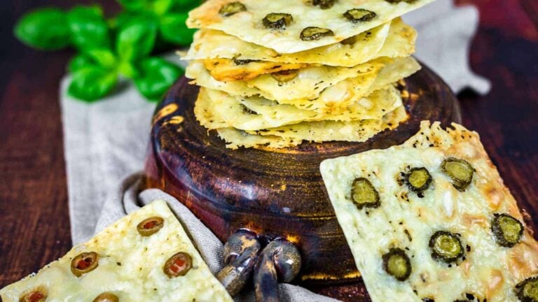 Square crackers with green olives are stacked on a round wooden board, with a few crackers arranged around it. Fresh basil leaves are in the background.