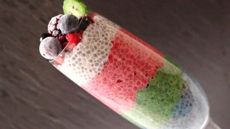 A glass filled with layered chia pudding in white, pink, and green colors, topped with frozen berries and a slice of kiwi.