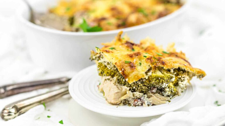 A slice of broccoli and cheese lasagna is served on a white plate, with a casserole dish and utensils in the background.