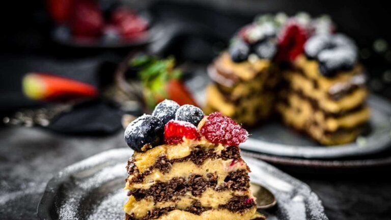 A slice of layered chocolate cake with cream, topped with blueberries, raspberries, and powdered sugar, sits on a plate with the rest of the cake in the background.