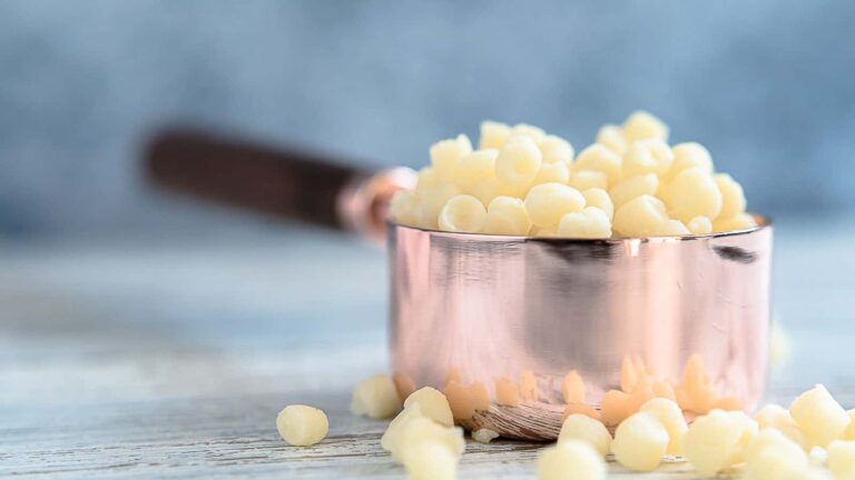 A copper measuring cup filled with white cocoa butter chips, with some chips scattered on a wooden surface.