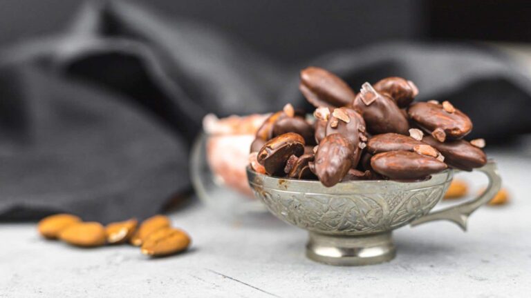 A silver cup filled with chocolate-covered almonds sits on a gray surface, with a few plain almonds scattered nearby and a black cloth in the background.