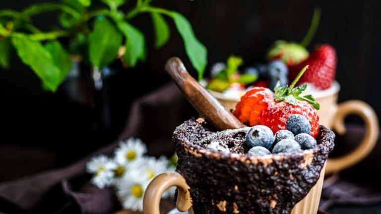 A mug filled with chocolate dessert, topped with blueberries, strawberries, powdered sugar, and a chocolate stick, with a blurred plant and flowers in the background.