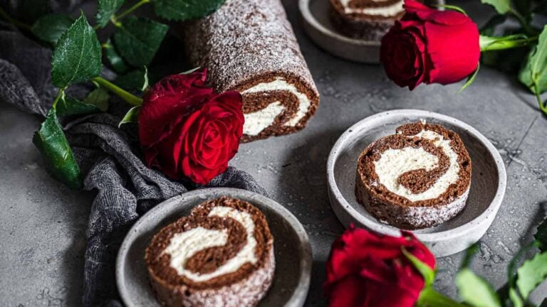 Chocolate Swiss roll cake with cream filling is sliced and served on small plates, surrounded by fresh red roses on a gray surface.