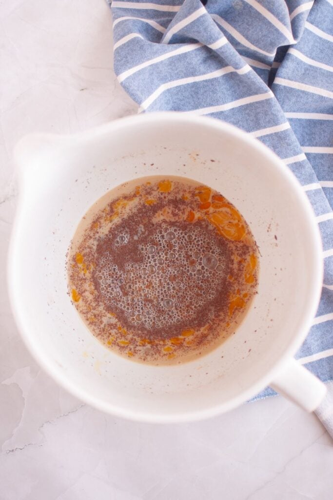 A white mixing bowl containing a mixture of wet ingredients, including eggs, milk, sugar, cinnamon, vanilla, and salt, sits on a white surface next to a blue and white striped cloth.