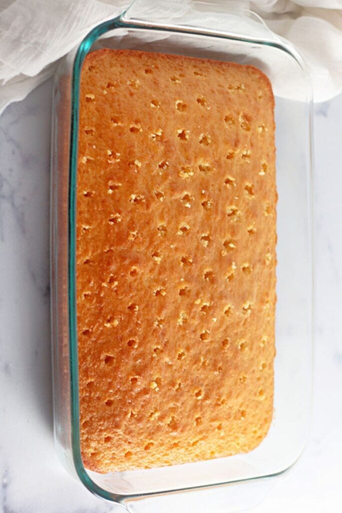 A rectangular yellow cake with evenly spaced holes on top, baked in a glass dish, sits on a white surface.