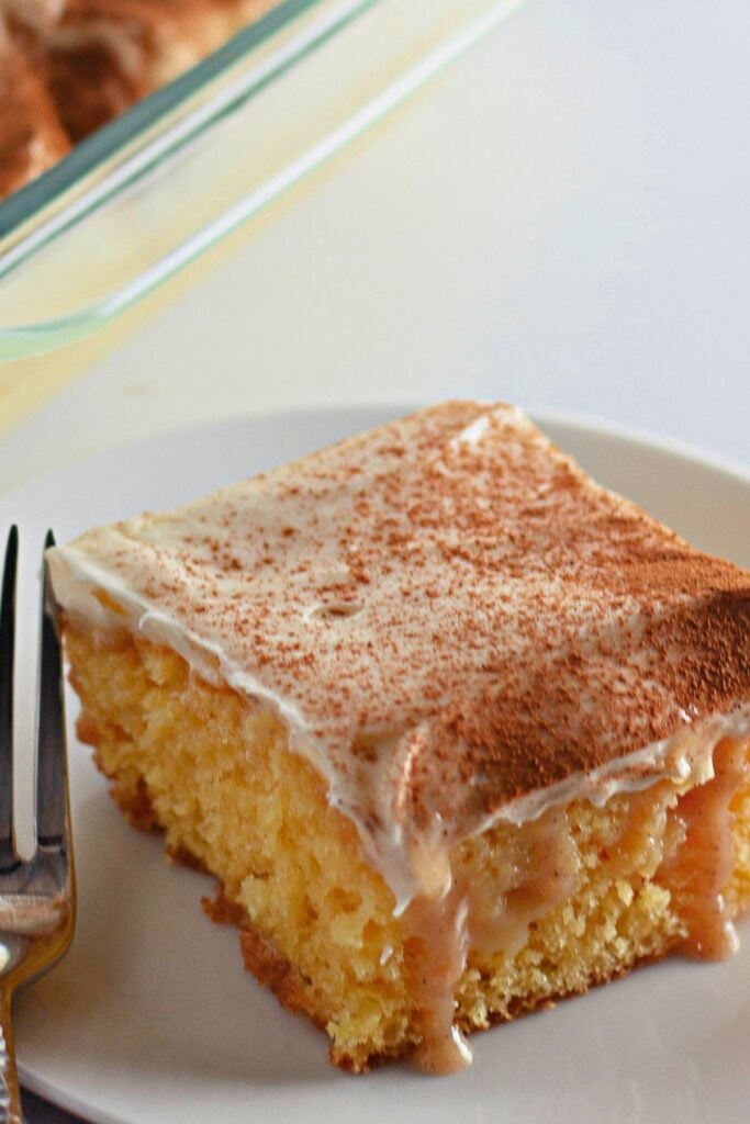 A square piece of moist yellow cake topped with creamy frosting and cinnamon, served on a white plate with a fork.