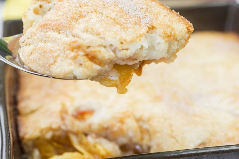 A close-up of a serving of peach cobbler being lifted from a baking dish, showing a golden, crumbly crust and peach filling.