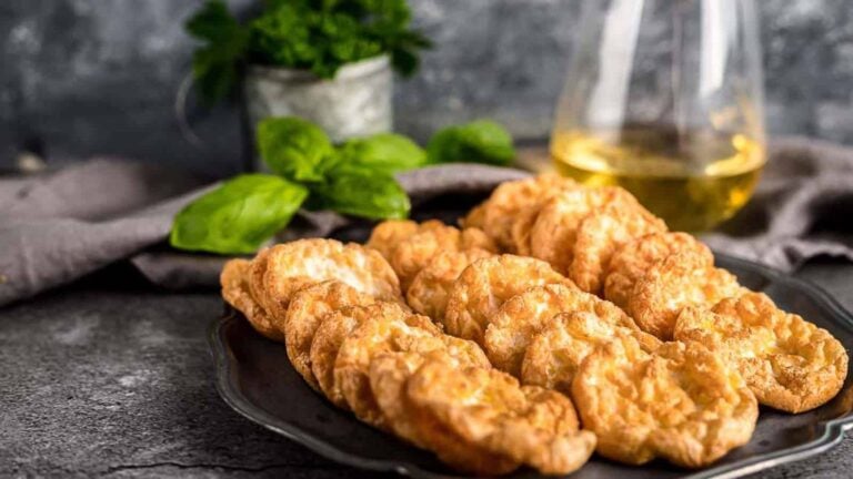 A dark plate of golden, crispy cloud bread slices is displayed on a stone surface, with fresh herbs and a glass jug in the blurred background.