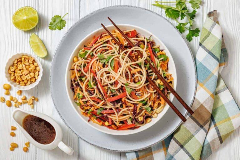 A bowl of noodle salad with vegetables, garnished with peanuts and cilantro, served with chopsticks, lime wedges, and soy-based sauce on the side.