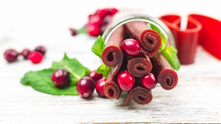 A glass jar with rolled fruit leather and fresh cranberries, garnished with mint leaves, on a white wooden surface.