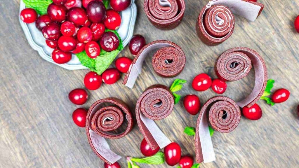 Several rolled pieces of fruit leather are arranged on a wooden surface, surrounded by fresh cranberries and a small plate of cranberries with mint leaves.