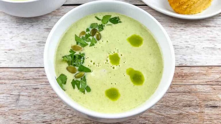 Bowl of green soup garnished with chopped herbs, seeds, and a drizzle of green oil, placed on a wooden surface.