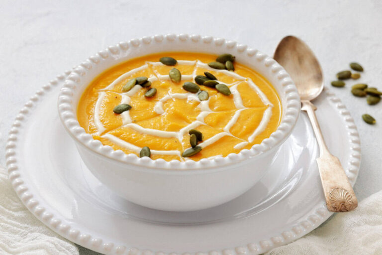 A bowl of pumpkin soup with a swirl of cream in a spiderweb pattern, topped with pumpkin seeds, served on a white plate with a spoon.