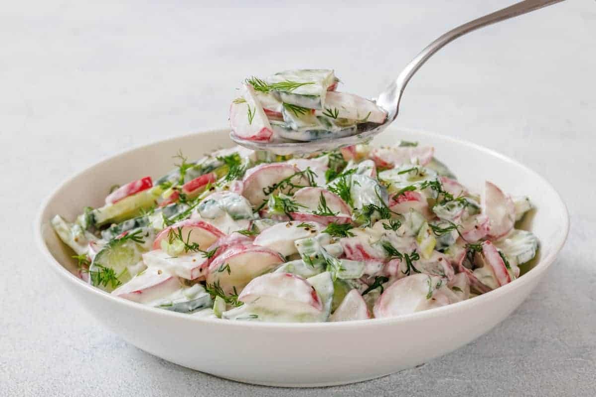 A bowl of Creamy Radish Salad garnished with dill, with a spoon lifting a portion above the bowl.