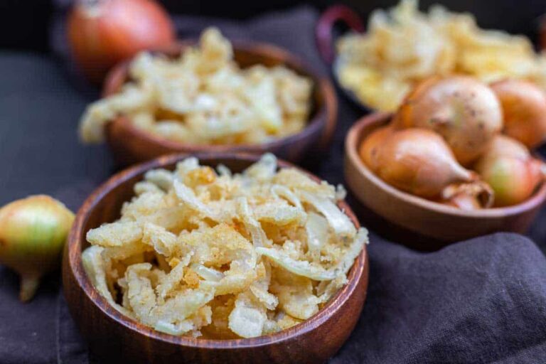 Three wooden bowls on a dark cloth, two filled with crispy fried onion strips and one with whole onions, with more onions and fried strips in the background.