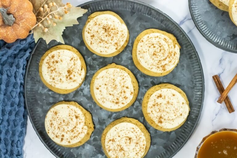 A round metal tray holds seven frosted cookies sprinkled with brown spice. Nearby are a small pumpkin, dried leaves, a blue textured cloth, and cinnamon sticks.