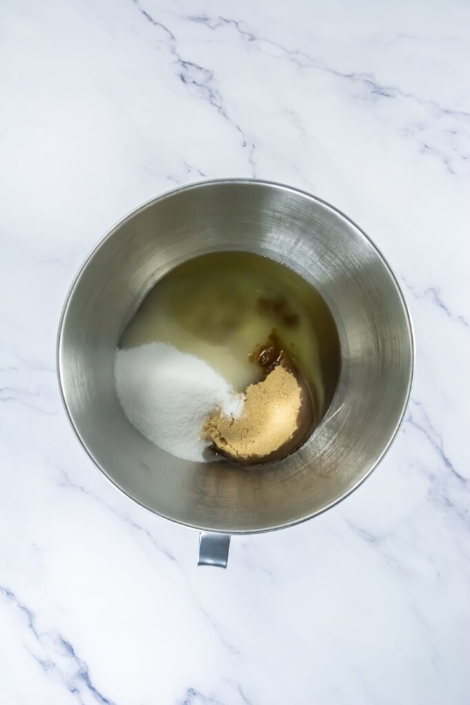 A metal mixing bowl contains sugar, brown sugar, and oil—perfect for creating Pumpkin Spice Whoopie Treats—on a white marble surface.