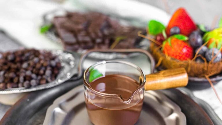 A small glass pitcher of melted chocolate on a plate, with a tray of dried fruit, a plate of chocolate pieces, and a basket of fresh berries in the background.