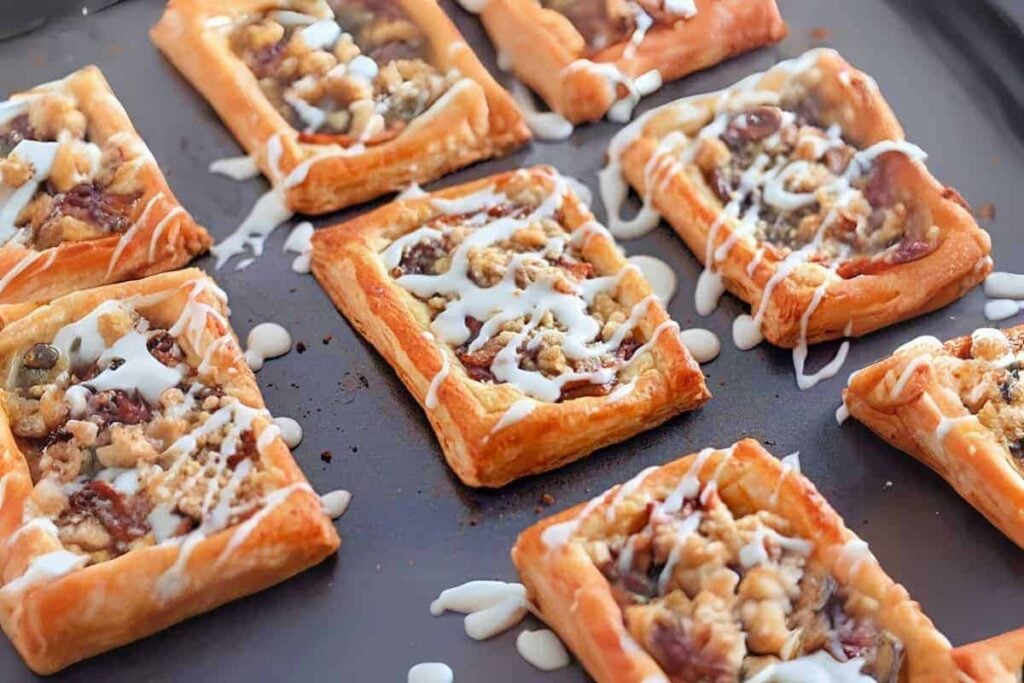 Rectangular puff pastries with a crumb topping and white icing drizzle are arranged on a dark baking tray.
